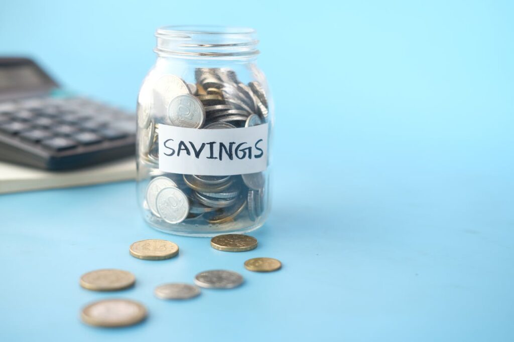 pexels-photo-9821387 Glass jar labeled 'Savings' filled with coins, beside a calculator on a blue background.